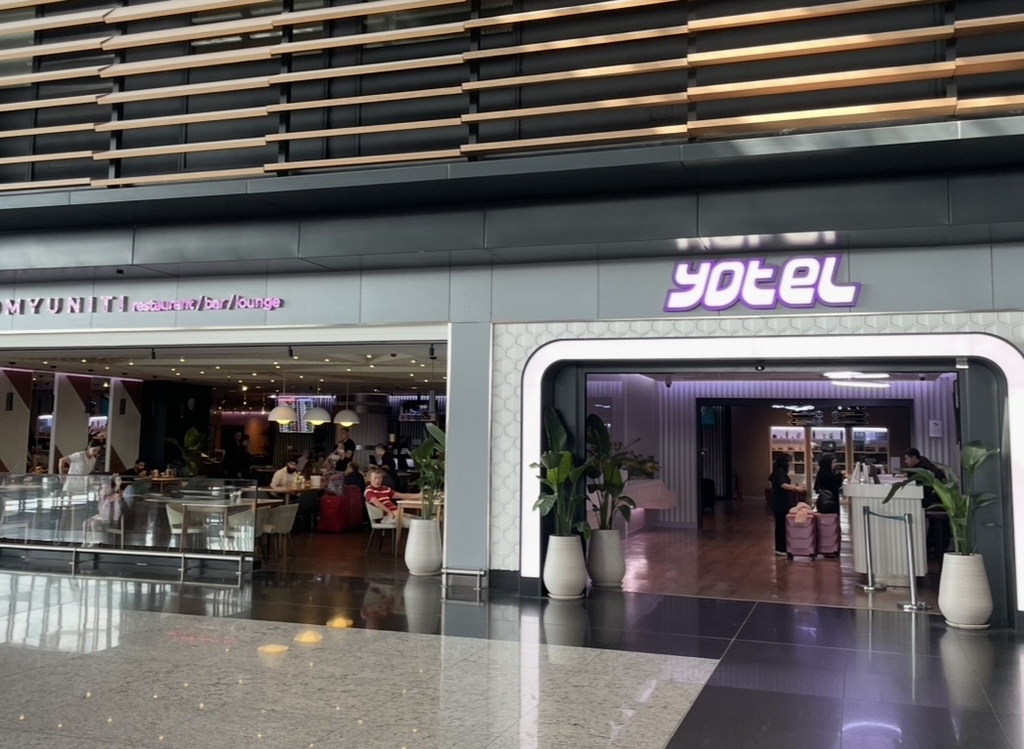 The entrance of YOTEL inside Istanbul Airport, featuring a futuristic design with white and purple lighting that stands out alongside the adjacent restaurant area, creating a comfortable space within the airport.
