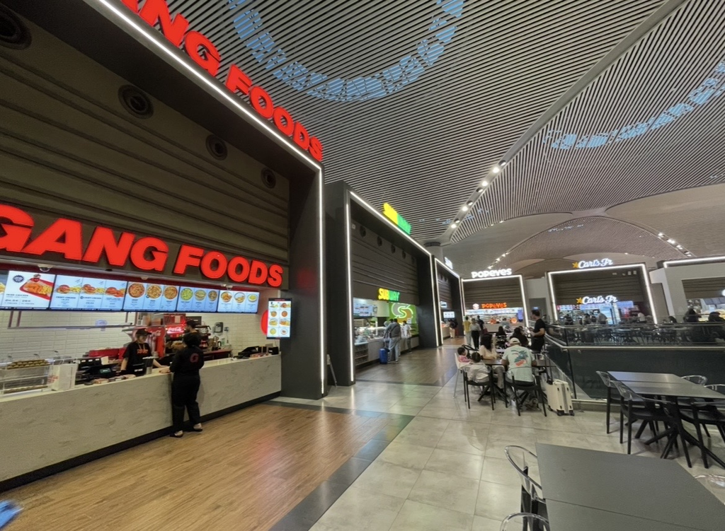 The food court at Istanbul Airport, a spacious area lined with multiple eateries including the red‑signed GANG FOODS, Subway, Popeyes, and Carl’s Jr., where travelers enjoy meals at tables in a lively atmosphere.