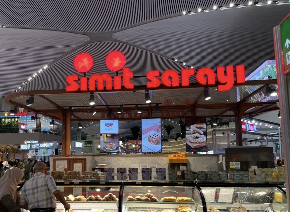 伊斯坦堡國際機場內的 Simit Sarayı，紅色招牌醒目的麵包店，玻璃櫃中陳列現烤的芝麻麵包與酥點，旅客在店內排隊點餐，氣氛熱鬧。