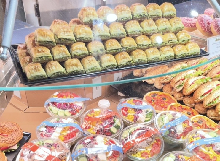 A tray of baklava displayed in a showcase, with golden layered pastry revealing bright pistachios, while the lower shelf features colorful salads and sandwiches in a well‑stocked food display.