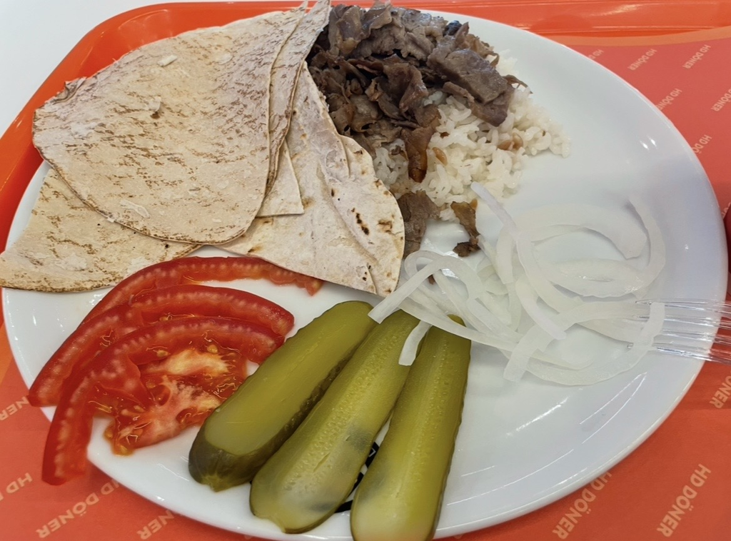 An HD Döner kebab set featuring juicy kebab meat served on thin flatbread, fluffy rice, and colorful tomatoes, pickles, and onions arranged neatly on a plate placed on an orange tray.