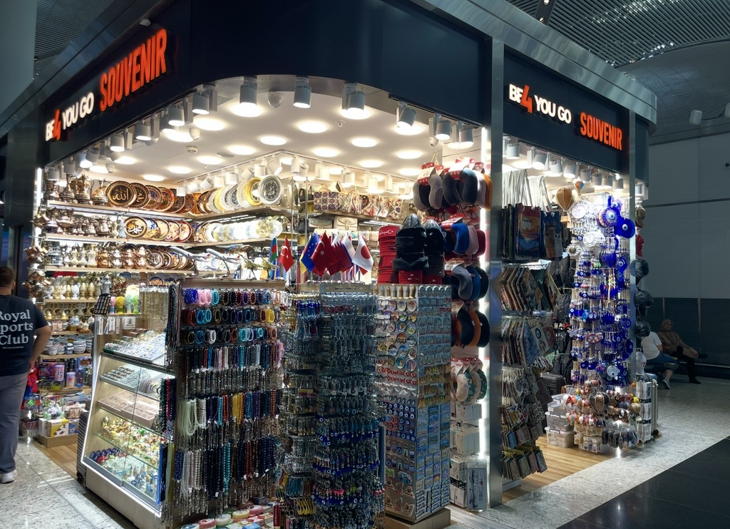 伊斯坦堡國際機場出發大廳的紀念品店，色彩鮮豔的盤子、茶壺、鑰匙圈、手鍊與土耳其避邪飾品等多樣商品陳列其中，旅客在熱鬧的店內駐足選購。