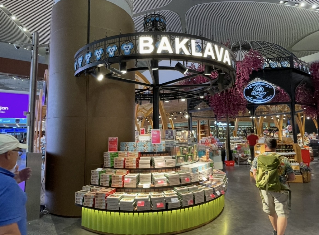 伊斯坦堡國際機場管制區內的紀念品店，中央以圓形堆疊的巴克拉瓦禮盒打造華麗展示，周圍有花卉裝飾與各式店舖，旅客在熱鬧空間中穿梭。