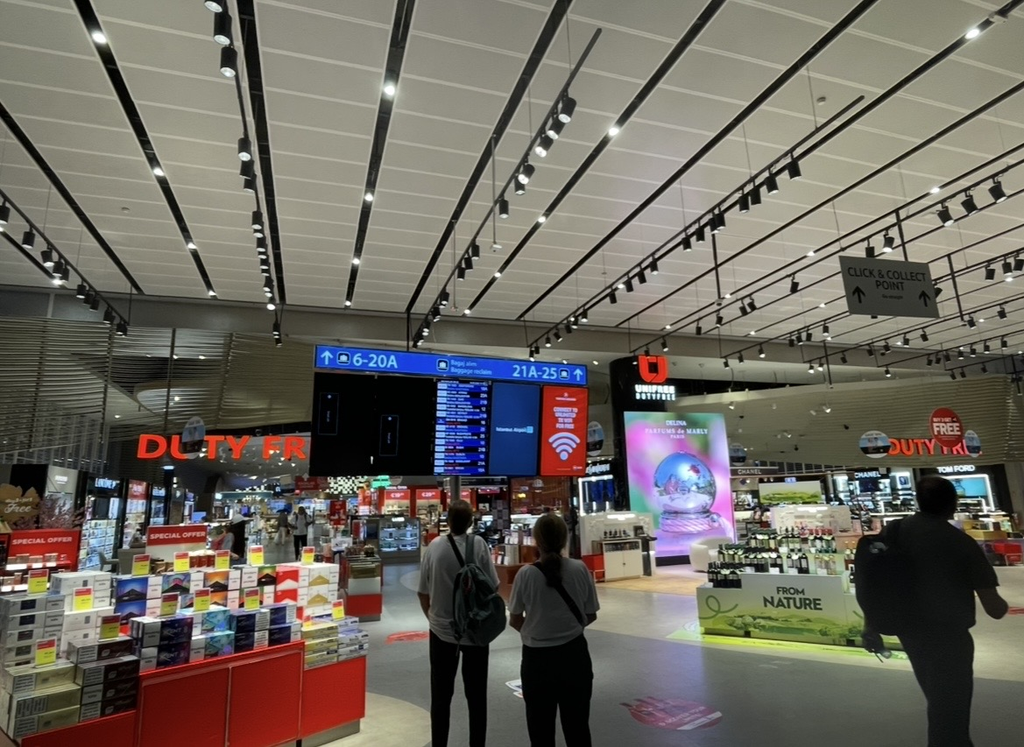 A duty‑free shop near the arrivals area of Istanbul Airport, centered around a large flight information board and surrounded by stores, creating a lively space where travelers check information while enjoying shopping.