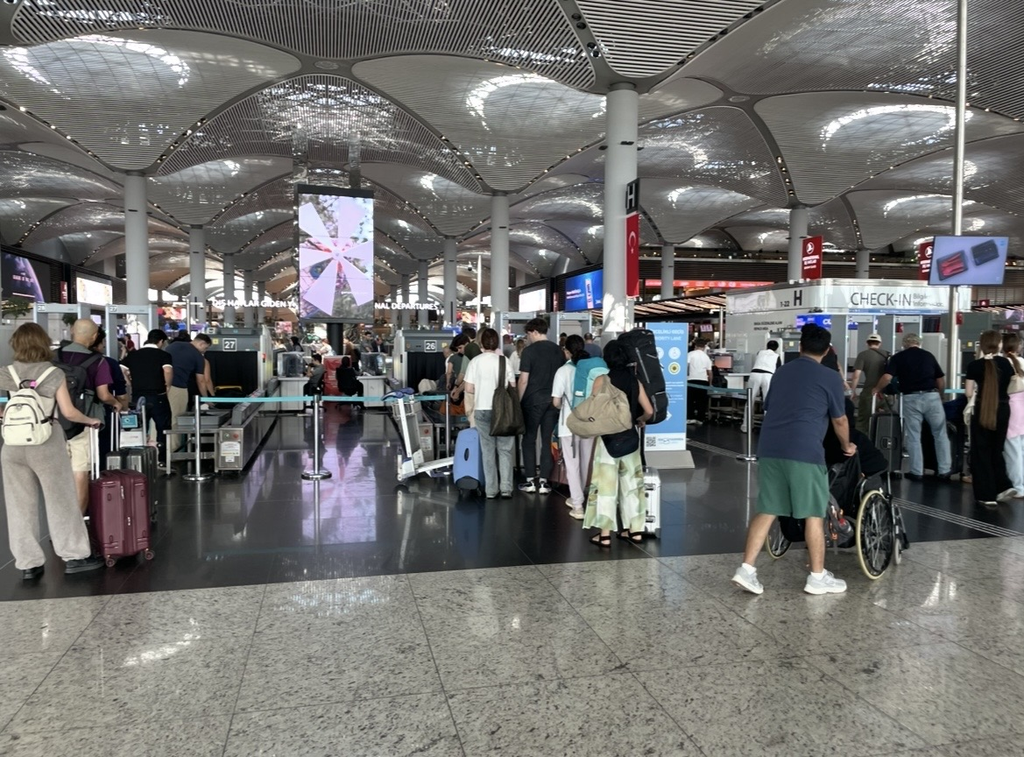 伊斯坦堡國際機場出發大廳入口的安檢區，旅客在報到櫃台前排隊，提著行李或使用輪椅的人在工作人員引導下前往檢查。