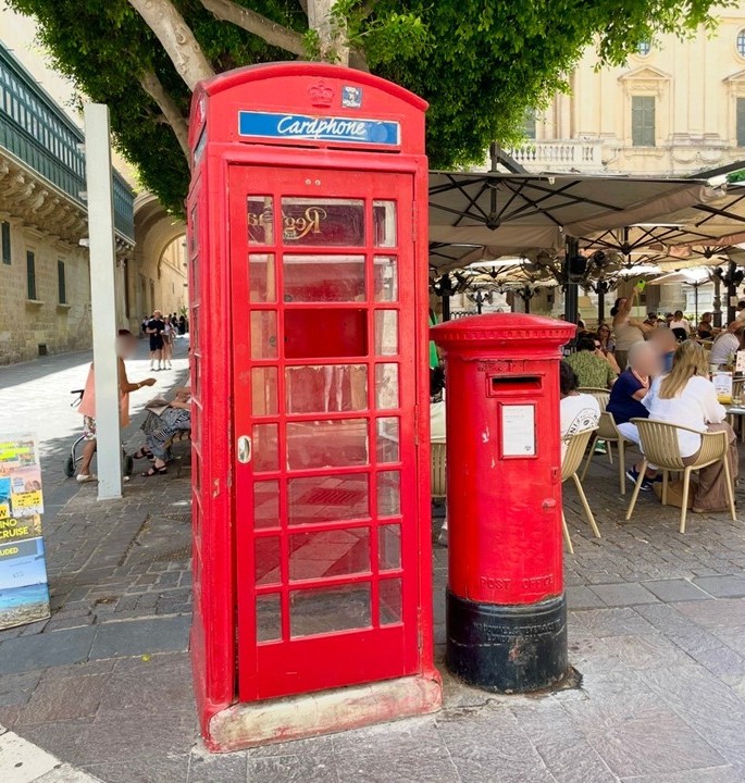 バレッタの街角に立つ赤い電話ボックスと郵便ポスト。イギリス統治時代の名残が残る歴史的な街並みで、カフェのテラス席や石造りの建物が並ぶ観光スポットの風景。