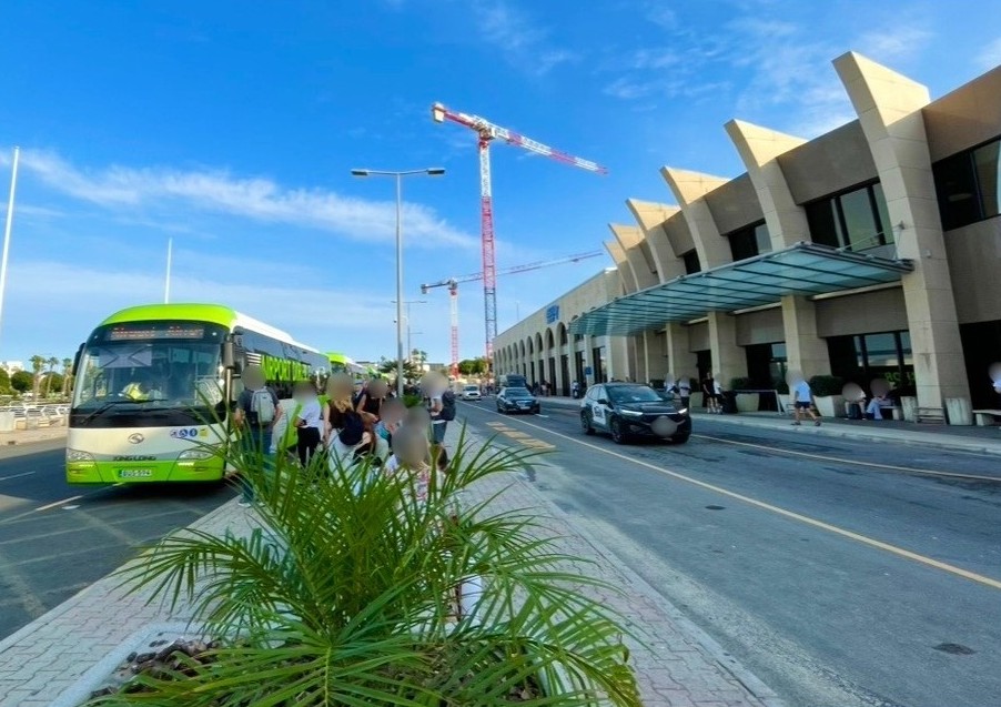 マルタ国際空港のターミナルビル外観と、道路を挟んですぐ向かい側の停留所に停車している緑と白の路線バスの風景。