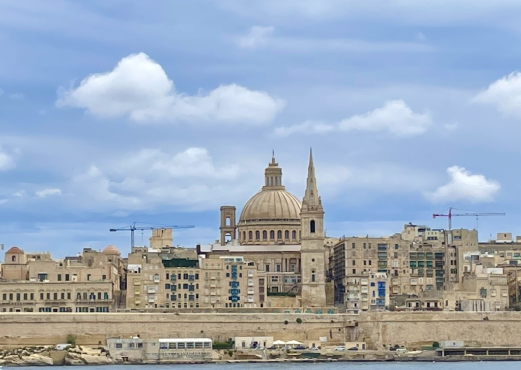 瓦勒他沿海延展的歷史街景，石造建築與醒目的圓頂並列，面向湛藍的地中海。柔和的天空色調與海岸線相互融合，呈現馬爾他的代表性風光。