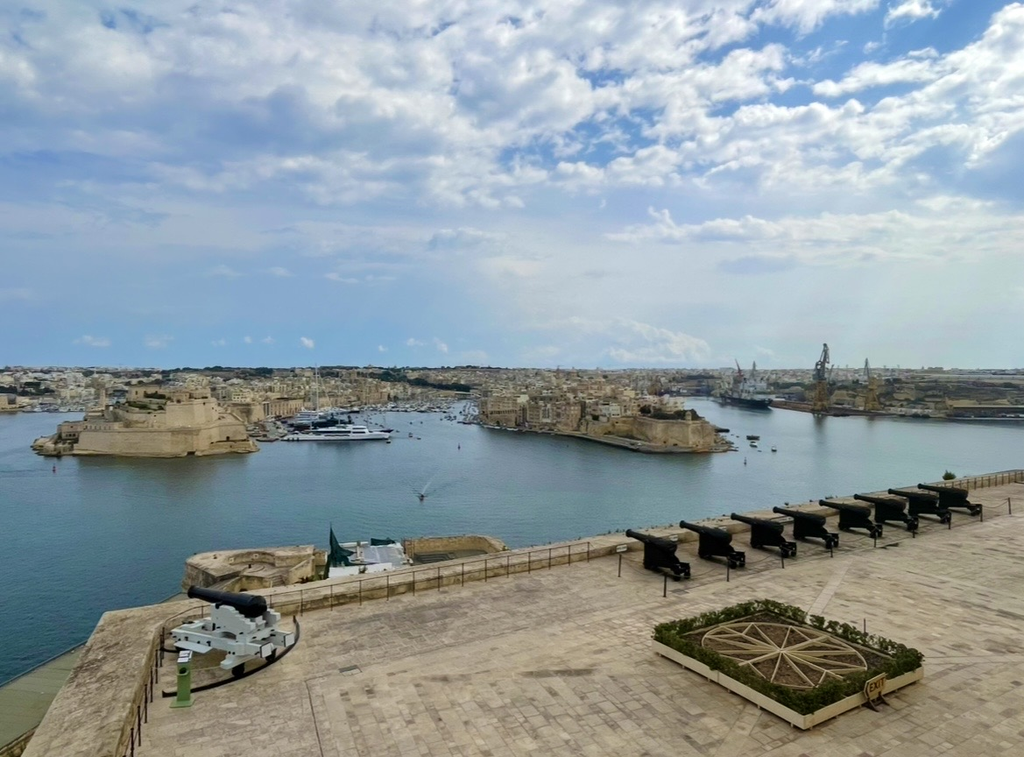 A panoramic view of the Grand Harbour from the Upper Barrakka Gardens, with aligned cannons, historic fortifications, and ships moving across the water creating a dramatic and iconic scene.