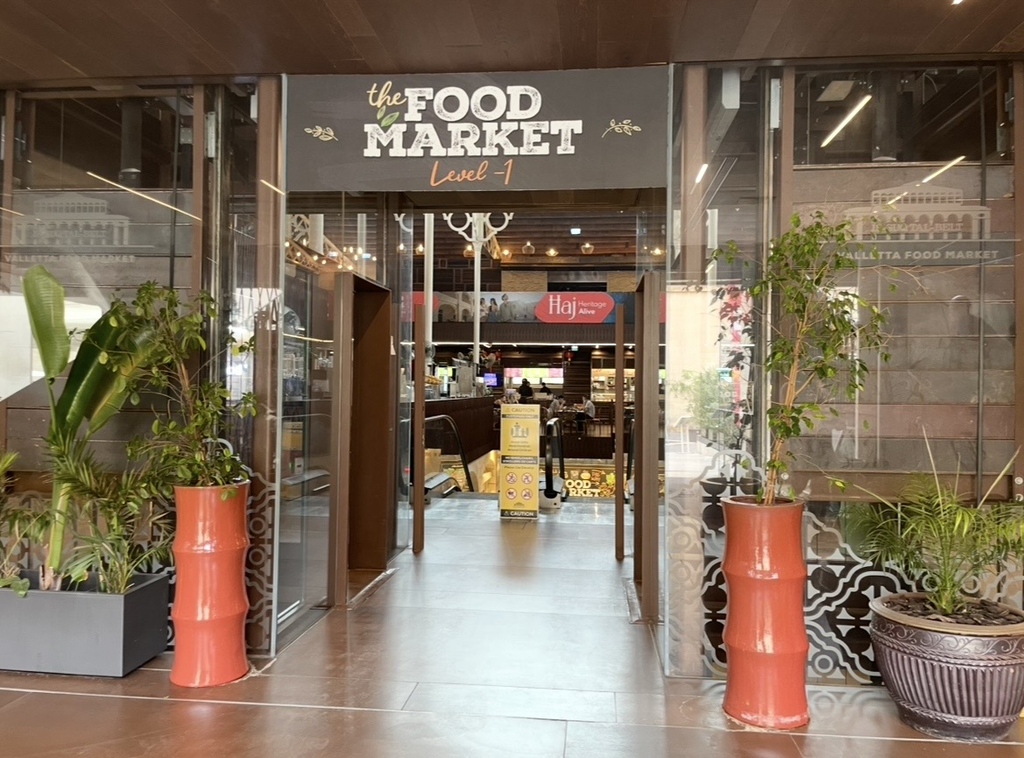 The entrance of Valletta’s Is‑Suq tal‑Belt food market, featuring a modern glass façade framed by greenery, leading into the renovated culinary complex.